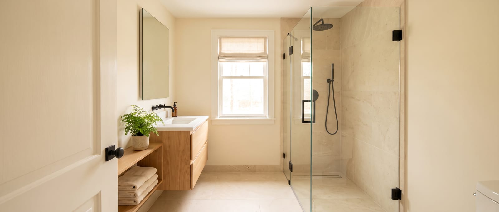 A compact bathroom with a corner glass shower, a wall-hung oak vanity and a window